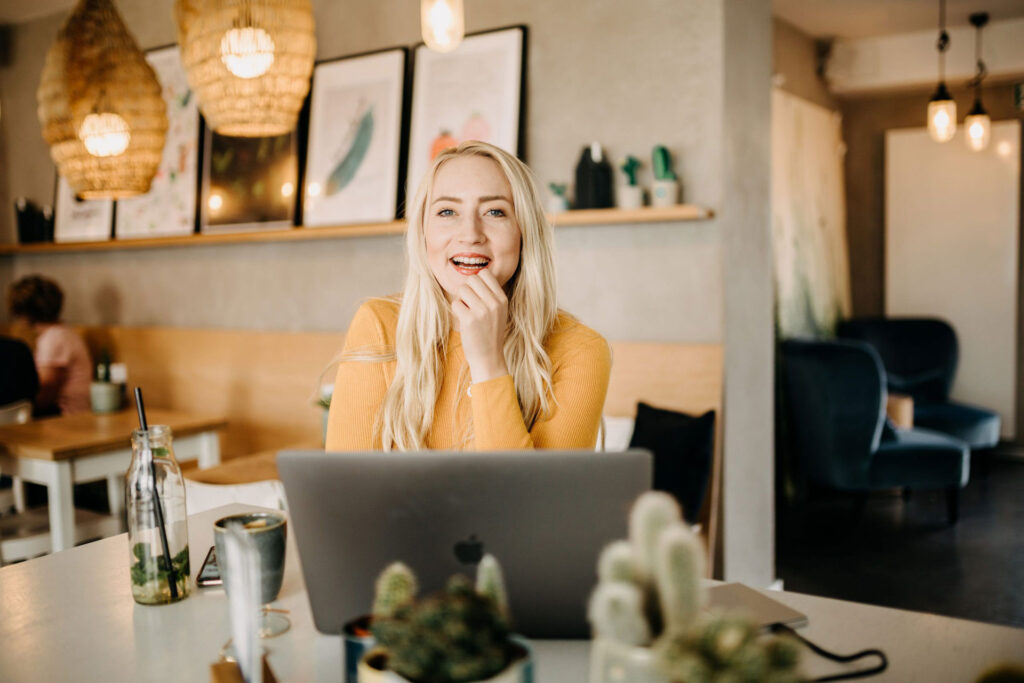 Jessica sitzt am Tisch im Cafe und schaut über ihr aufgeklapptes Notebook in die Kamera